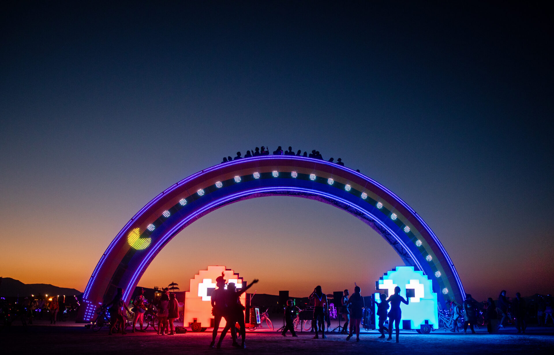 People stand on top of and dance under the Rainbow Bridge with mutant vehicles shaped like Pacman ghosts at sunset.