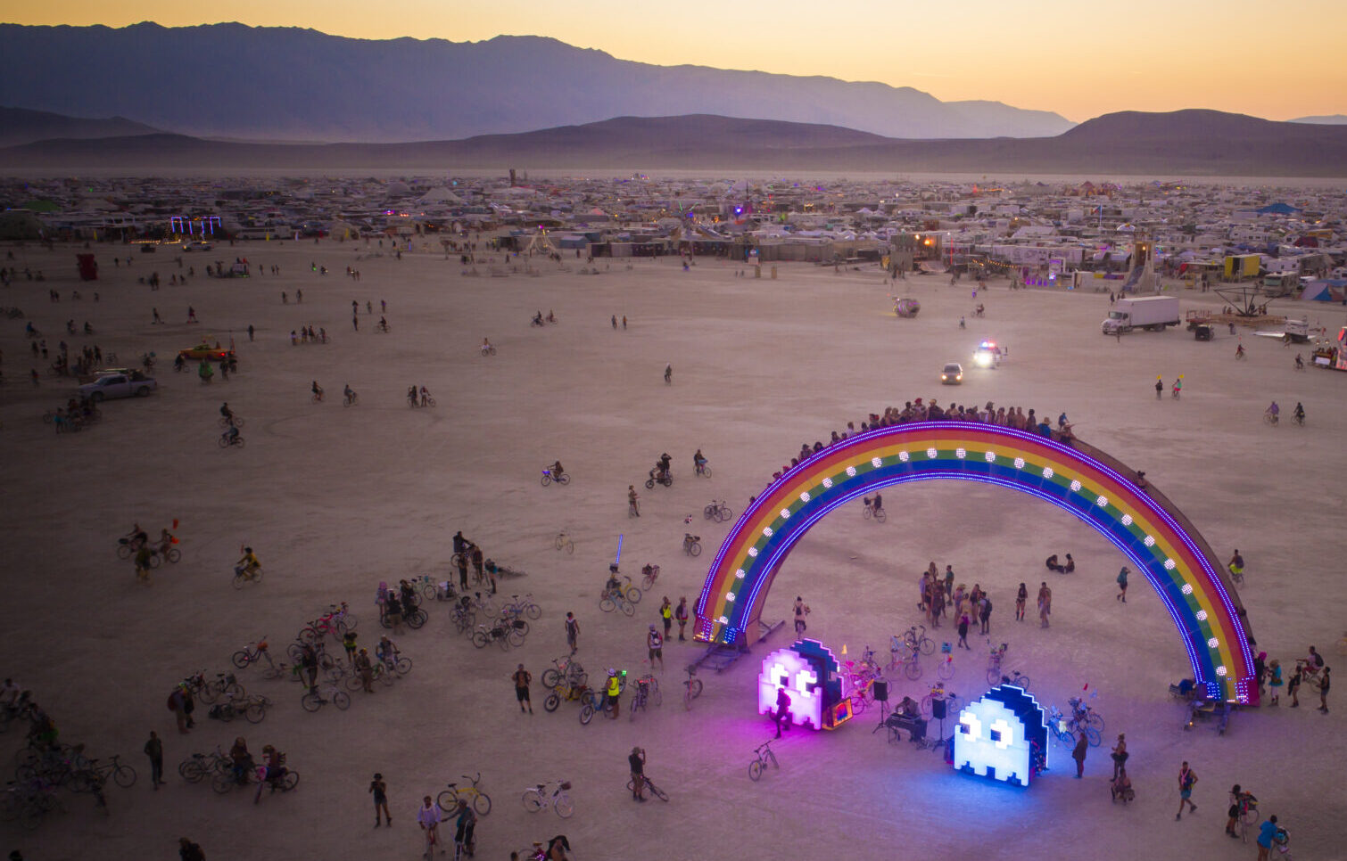 At sunset, two illuminated pac-man ghosts under a rainbow bridge are surrounded by people frolicking.