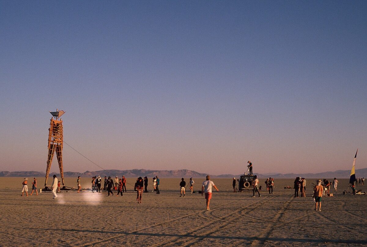 Wooden sculpture of a man, standing on the playa with people mingling nearby.