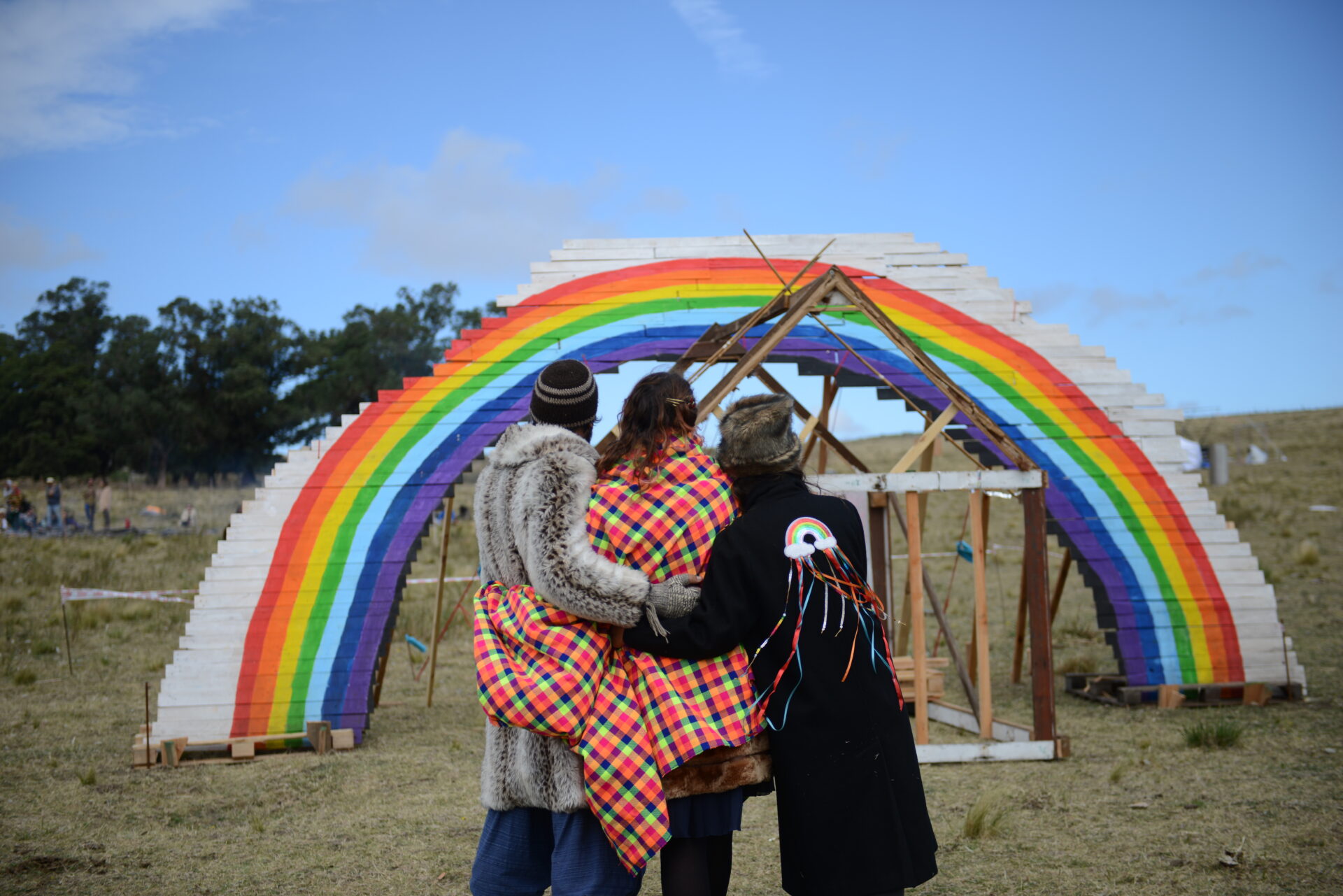 Three people stand in front of a rainbow sculpture. 